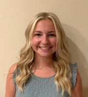 The image depicts a person with long, wavy blonde hair, smiling at the camera. They are wearing a light blue, textured top with ruffled sleeves. The background is a plain, light-colored wall, which provides a neutral backdrop to the subject.