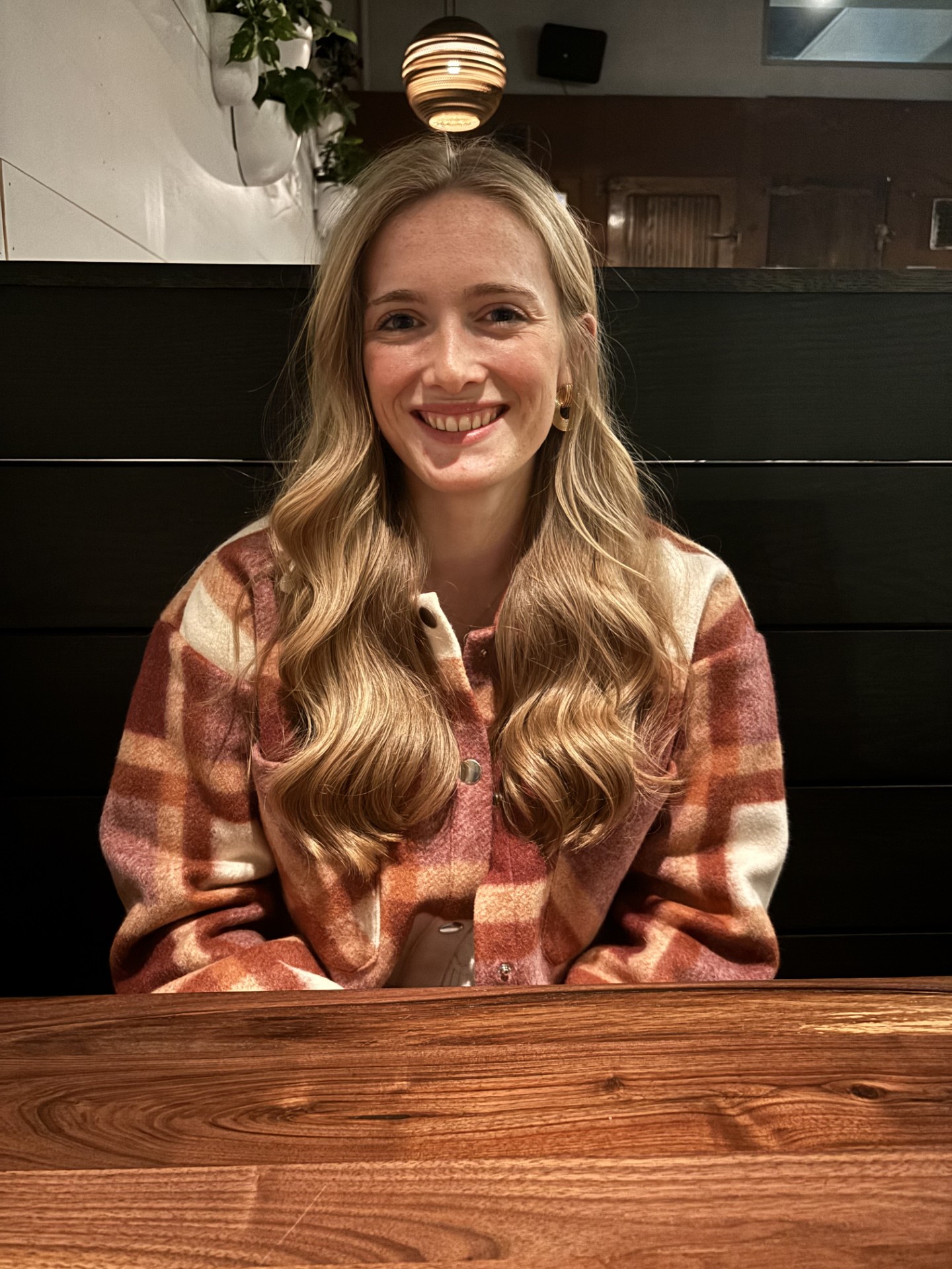 The image features a person sitting at a wooden table in what appears to be a cozy, dimly lit restaurant. The person is wearing a plaid jacket with shades of red, orange, and white. They have long, wavy blonde hair and are smiling at the camera. Behind them is a dark wall with a spherical pendant light hanging above, providing warm lighting. Some potted plants are visible on the left side against a white wall.