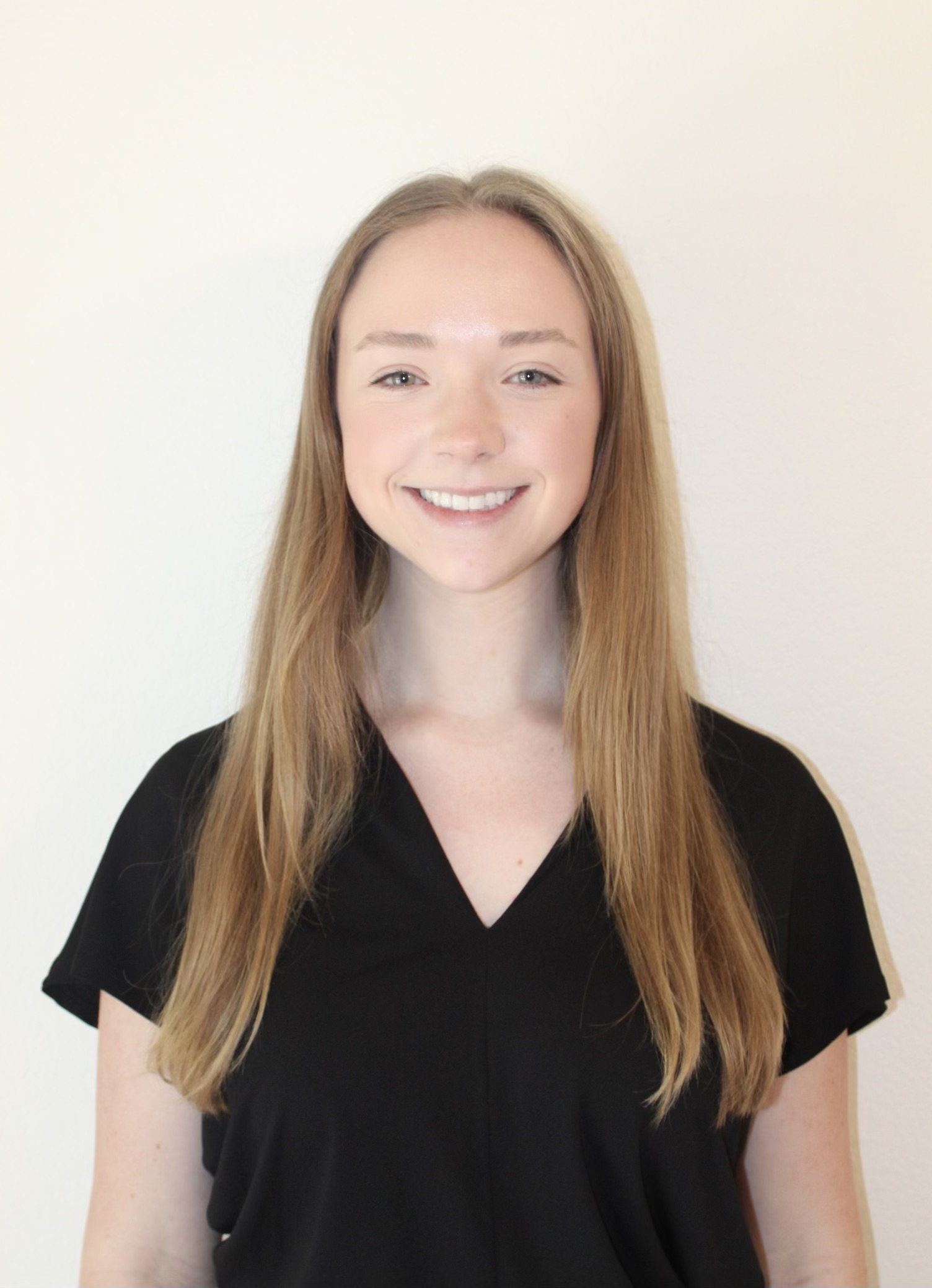 The image shows a person with long, straight hair standing against a plain, light-colored background. The person is wearing a black, short-sleeved shirt and is smiling directly at the camera. The hair is parted in the middle and falls down over the shoulders. The lighting is even, highlighting the person's facial features and giving the image a soft, natural appearance.