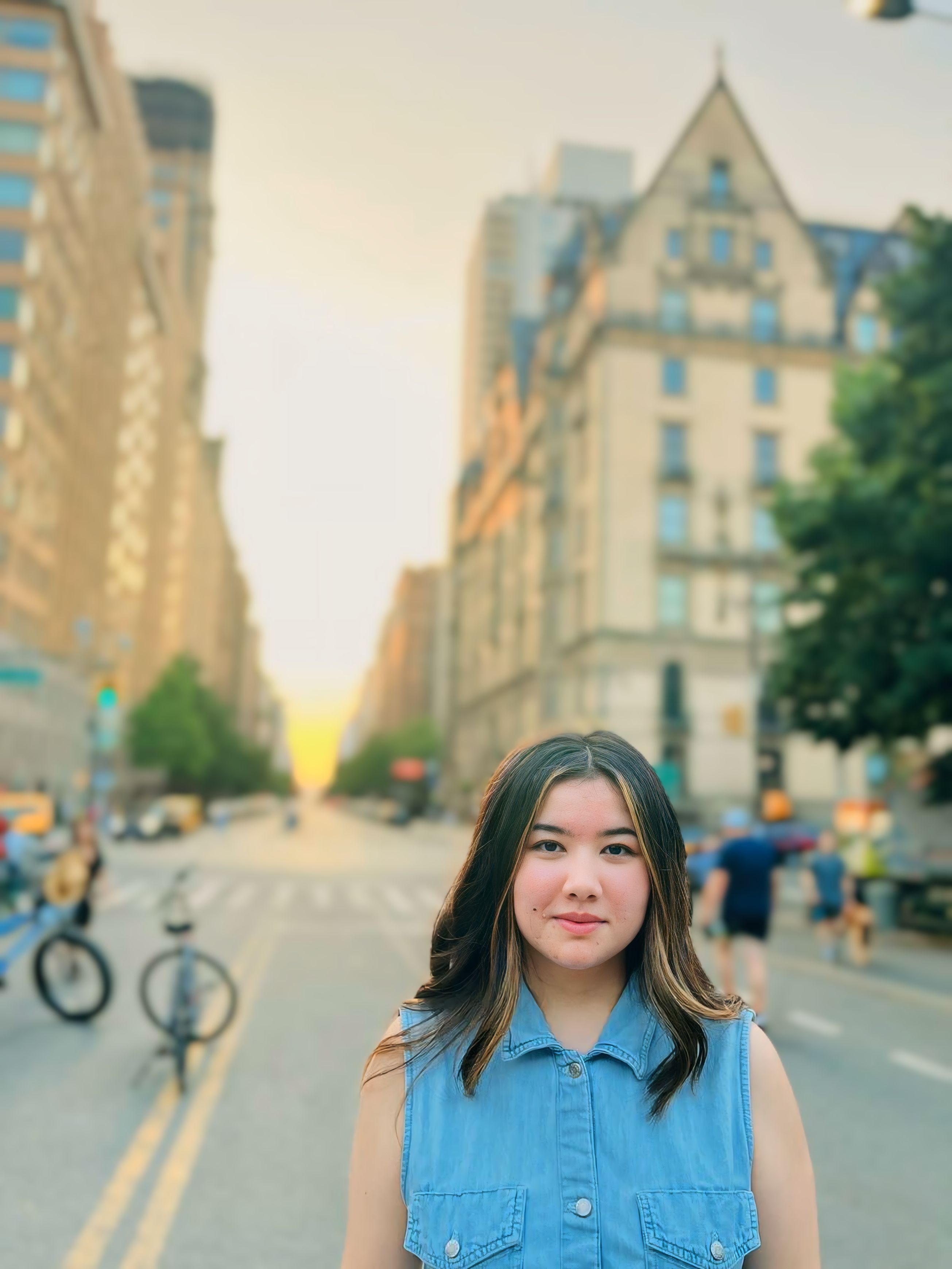 A young woman in a denim top standing on a city street at sunset with tall buildings in the background.