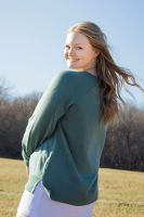 The image shows a person standing in a grassy field under a clear blue sky, captured from a three-quarter angle facing left. They're wearing a long-sleeved, teal sweater and light-colored pants. Their long hair is loose and gently blown by the wind. A line of trees is visible in the distant background.