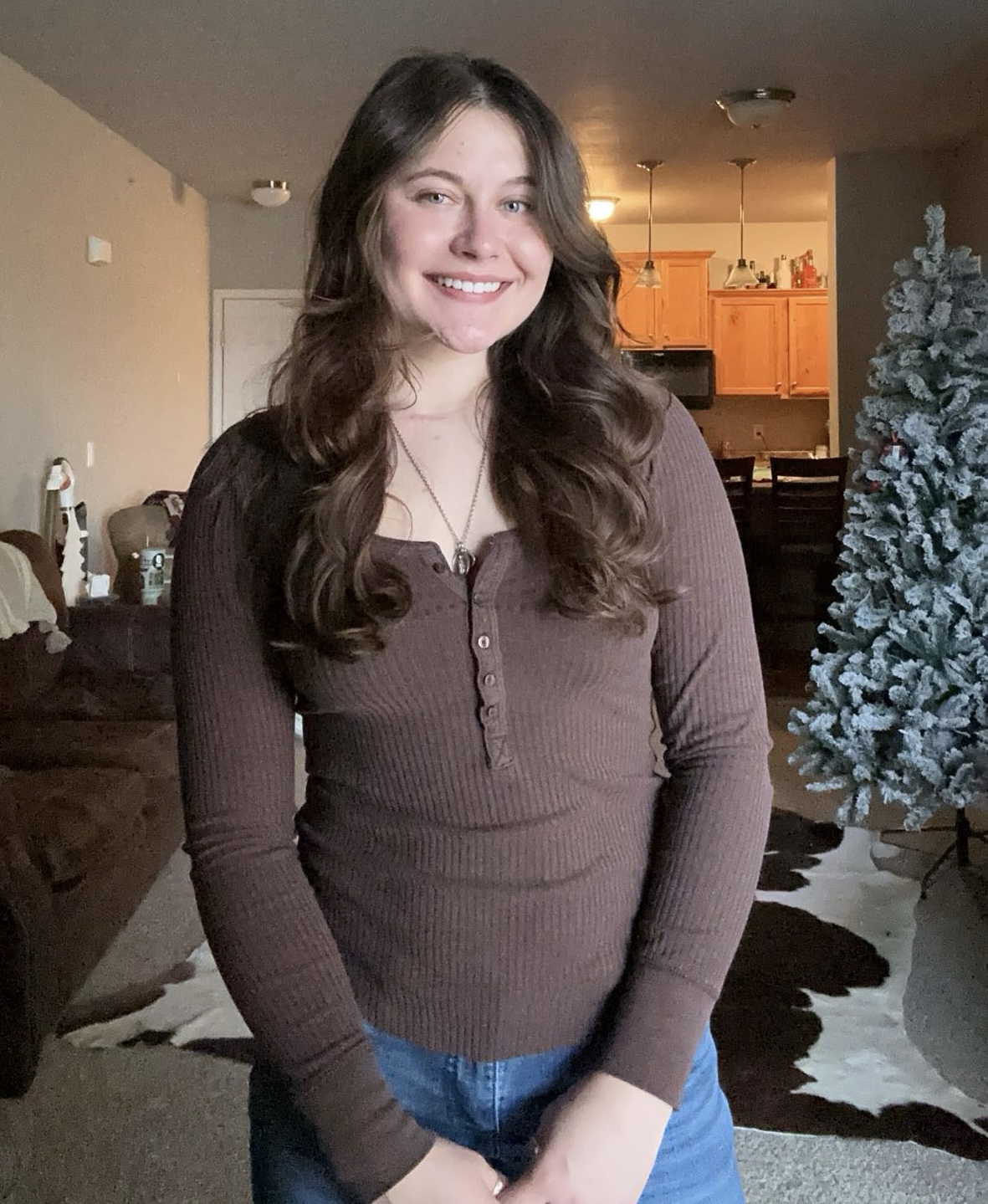 A woman in a brown long-sleeve shirt and jeans stands indoors, smiling. There is a decorated Christmas tree and a cowhide rug in the background.