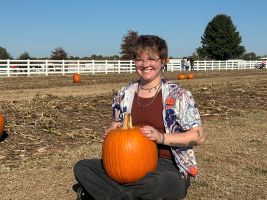 The image shows a person sitting on a patch of dry grass and dirt, holding a bright orange pumpkin. The individual is smiling, wearing glasses, and dressed in a colorful patterned shirt with a reddish-brown t-shirt underneath. The background features several scattered pumpkins, a white wooden fence, and a clear blue sky. In the distance, there are a few trees and a group of people walking or standing near the fence.