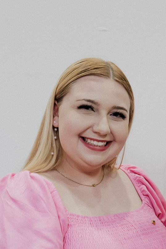 The image features a person with long blonde hair, smiling and looking at the camera. The individual is wearing a bright pink dress with puffed sleeves and a shirred bodice. They have pearl earrings and a delicate necklace. The background is plain white, ensuring the person is the main focus of the image.