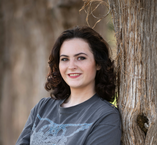 Person with curly hair leaning against a tree trunk, smiling in an outdoor setting.