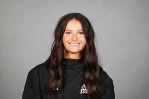 The image shows a woman with long, wavy brown hair smiling at the camera. She is wearing a dark jacket with the logo "Arizona State" visible on the front. The background is a plain, light gray color, giving a neutral backdrop that highlights the subject.