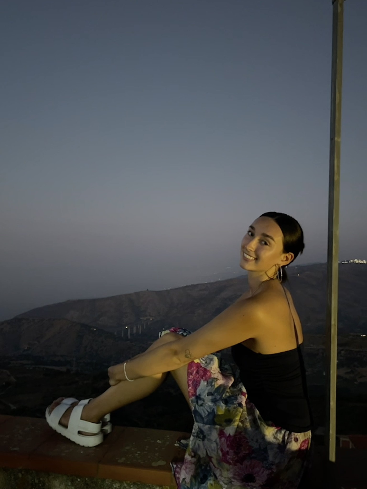 Woman is sitting on a ledge at dusk with mountains in the background.